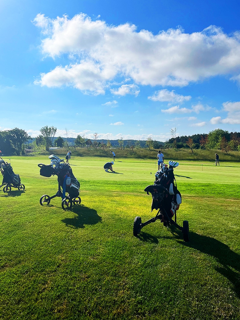 Drei Golfbags im Vordergrund dahinter mehrere junge Golfspieler auf einem Übungsgrün